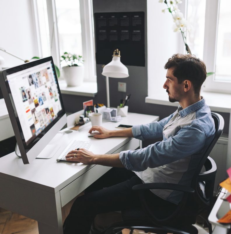 Man seated at a white desk using a large desktop monitor in a bright office space.